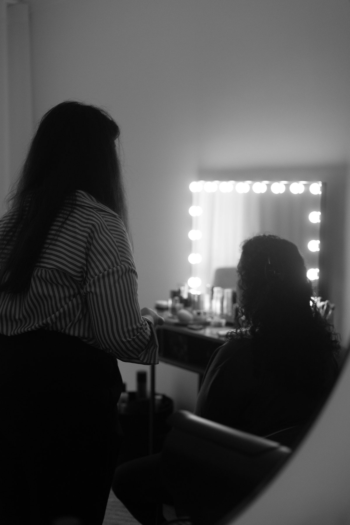 bastidores de maquilhagem para sessão fotográfica, duas mulheres de costas, uma a maquilhar e outra a ser maquilhada, no ambiente vê-se um espelho com luzes e um móvel com várias maquilhagens