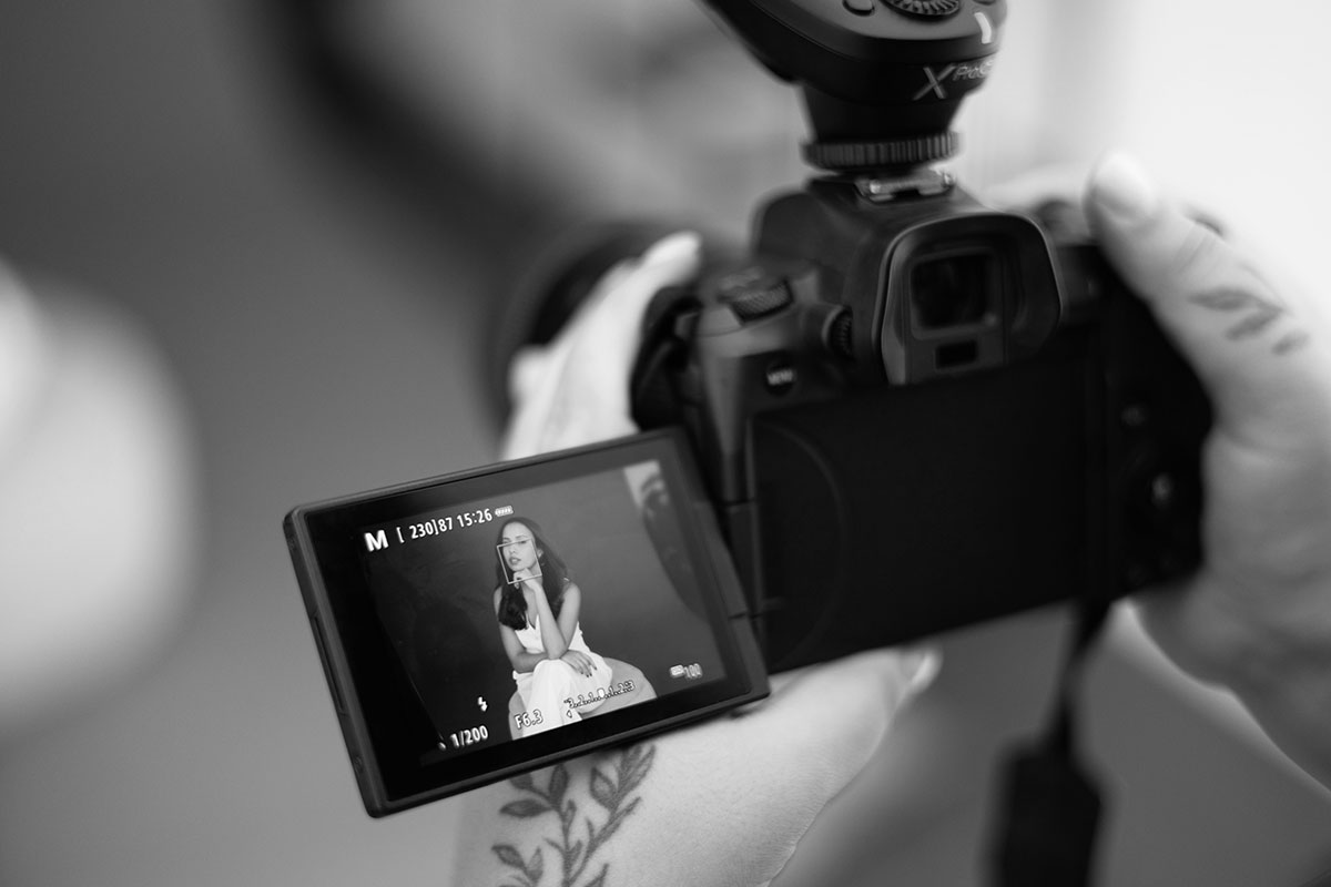 câmera fotográfica nas mãos da fotógrafa, no visor uma mulher sentada num puff, com a mão ao queixo e vestida de branco