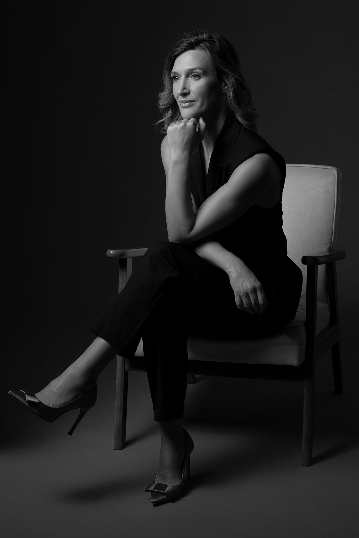 Retrato feminino editorial em estúdio, em preto e branco, sentada em cadeira com look total black e expressão confiante