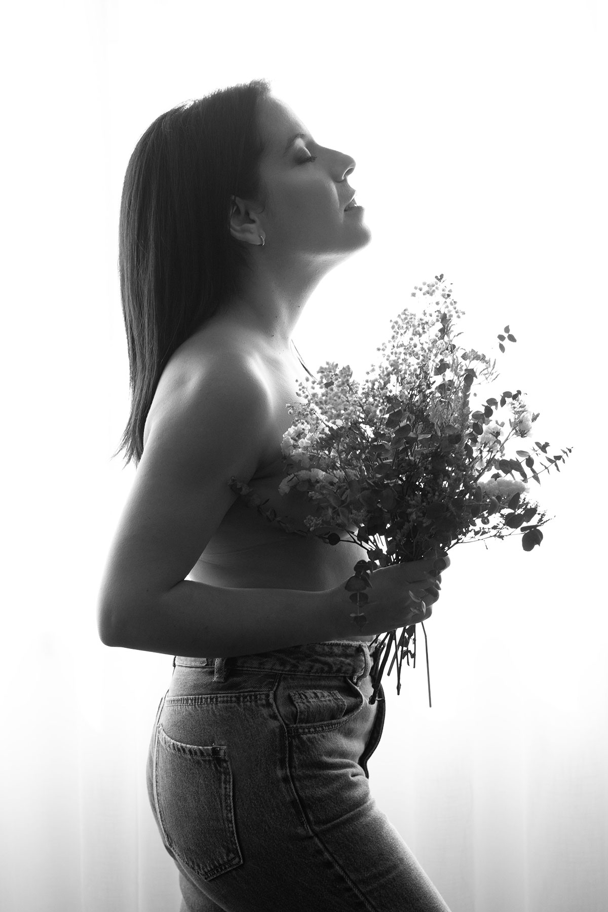 retrato em contra-luz, de uma mulher de perfil em tronco nu, a segurar flores para tapar, em baixo usa calças de ganga